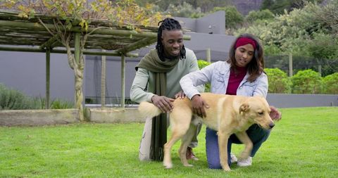 Diverse couple crouching petting golden-coated dog on lawn near pergola in garden
