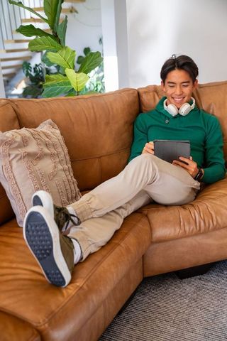 Asian man relaxing on tan leather sofa using tablet with headphones in modern living room
