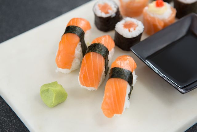 Freshly prepared sushi including salmon nigiri and maki rolls displayed on a white plate with a piece of wasabi and a small dish for soy sauce. Ideal for use in articles, blogs, and menus featuring Japanese cuisine, recipes, and dining experiences.