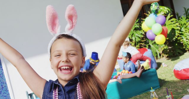 This image shows a young girl wearing bunny ears, smiling brightly at an outdoor birthday party. In the background, other children are playing and having fun. Balloons and decorations add to the festive atmosphere. Perfect for websites or advertisements related to children's events, party supplies, or family activities.