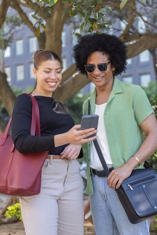 African american friends sharing smartphone under tree canopy in urban park, smiling