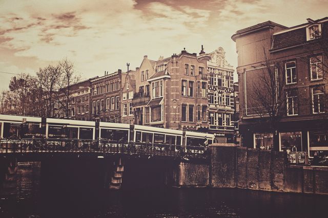 This image showcases a vintage urban cityscape with historic buildings lining a canal and a bridge crossing over the water. The sepia tone adds a nostalgic feel, reminiscent of old times in a European city. This can be used for travel blogs, historical articles, architectural features, postcards, backgrounds, or any project evoking nostalgia and old-world charm.
