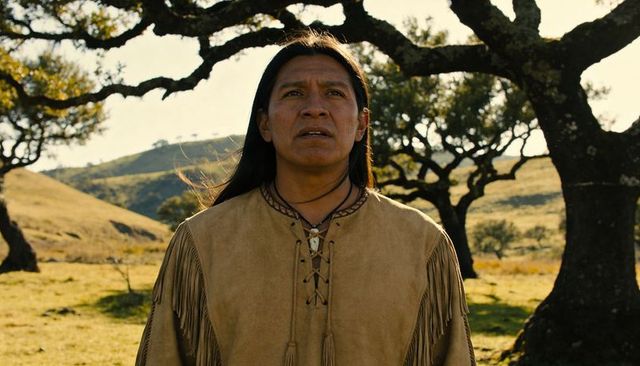 Native american man standing in sunlit meadow wearing fringed suede tunic and pendant