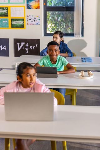 Smiling boy using tablet while classmates working on laptops in diverse classroom