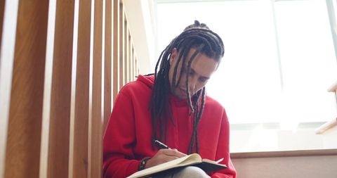 African american man with dreadlocks writing in journal on stair landing by backlit window
