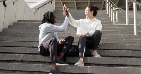 Friends high-fiving on urban stairs sharing pastry and backpack in sunlit city