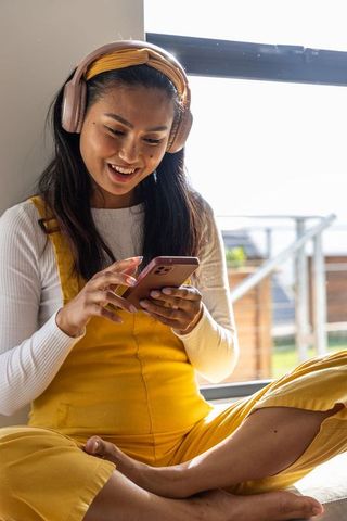 Young asian woman in mustard overalls with pink headphones smiling while using smartphone
