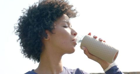 Young woman drinking from reusable bottle outdoors, fitness hydration, curly hair closeup