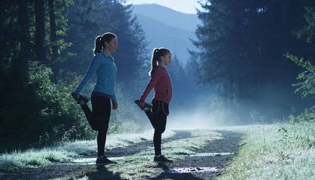 Mother and daughter stretching on misty forest trail at dawn in athletic jackets