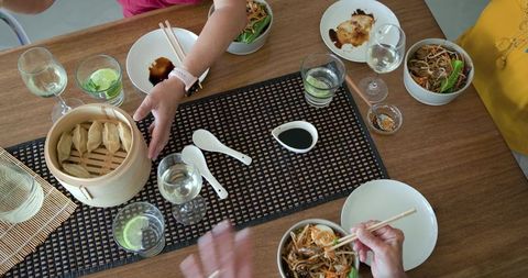 Female friends reaching across wooden table sharing bamboo-steamed dumplings and noodles