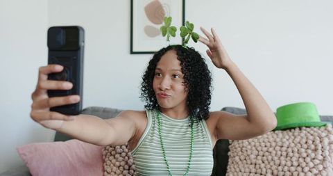 African american woman taking selfie wearing shamrock headband and green beads on cozy couch