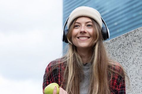 Smiling woman listening to music with headphones holding green apple in urban scene