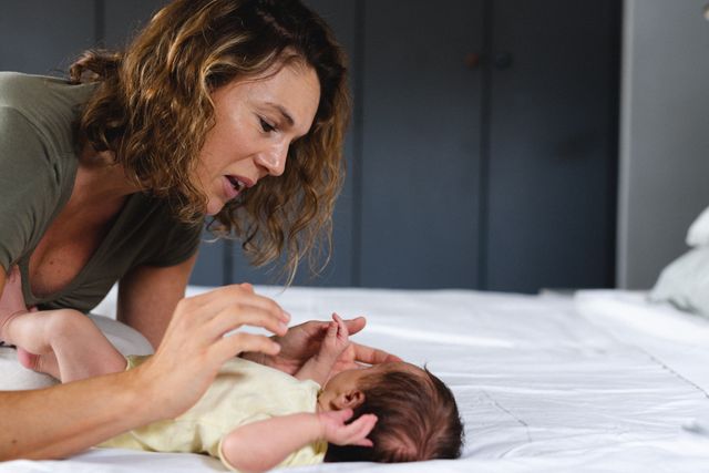 Mid adult Caucasian mother engaging with her newborn baby on a bed in a cozy bedroom. Ideal for use in parenting blogs, family care articles, and advertisements for baby products. Highlights themes of family bonding, maternal love, and early childhood development.