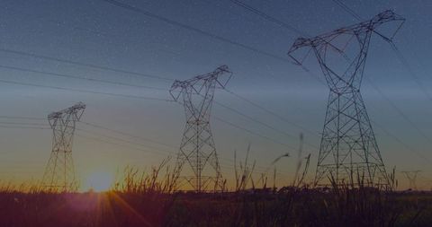 Silhouetting high-voltage transmission towers at sunset over rural field with tall grasses