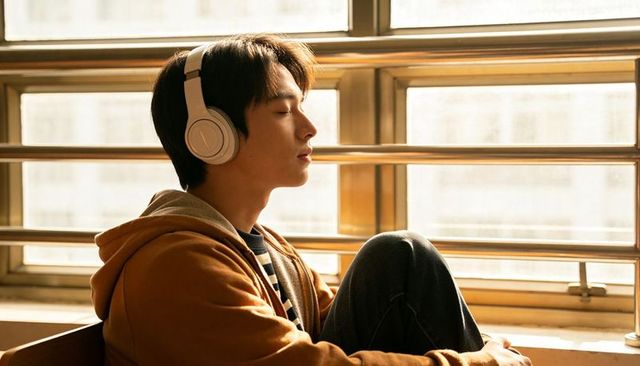 Young asian man listening to music on headphones, sunlit window bench, peaceful moment