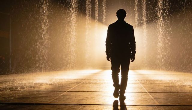 Silhouetted man walking through backlit fountain at night with golden water curtain