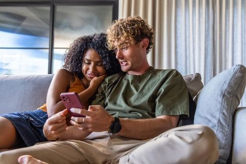 Interracial couple relaxing on sofa sharing smartphone, cozy living room connection