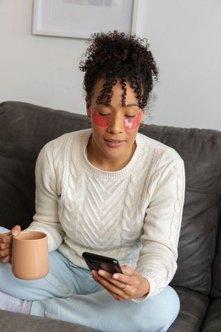 African american woman sitting on couch holding mug and smartphone with under-eye patches