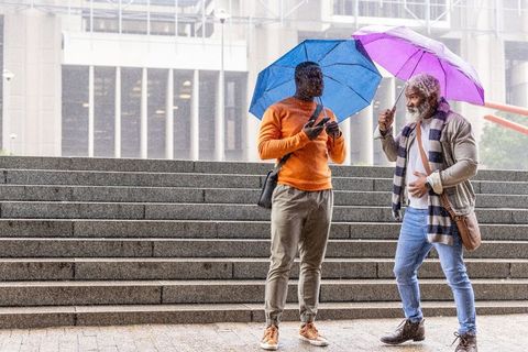 Two african american men holding umbrellas chatting and checking phone on city steps in rain