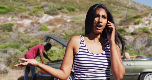 Woman wearing sleeveless striped top on phone while standing next to car with hood up. Ideal for content relating to road assistance services, car problems, mobile phone communication, travel issues, emergency help, auto repair services. Captures the essence of a rural area.