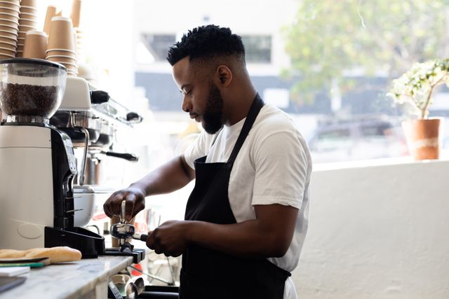 Young African American barista preparing coffee using an espresso machine in a modern cafe. Ideal for illustrating concepts related to cafe culture, small businesses, hospitality industry, and professional barista skills. Can be used in articles, blogs, and promotional materials for coffee shops and barista training programs.