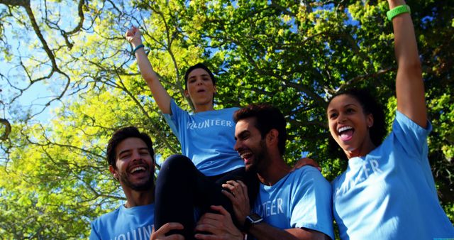 Group of volunteers wearing blue shirts, cheering and raising hands outdoors with trees in the background. Great for use in campaigns promoting volunteerism, teamwork, community service, and charity events. Ideal for illustrating concepts of unity, support, and group success.