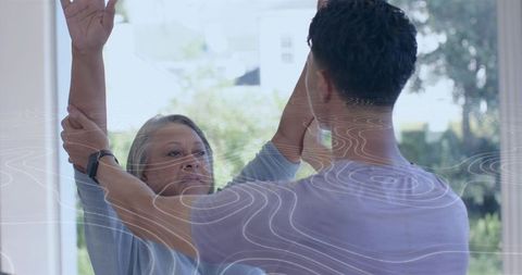Caregiver guiding senior through arm raising exercise near sunlit window home care smartwatch
