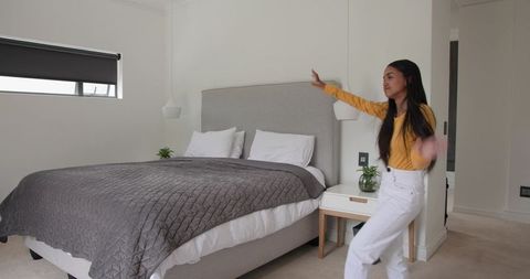 Woman late 20s showing modern minimal bedroom, gesturing toward bed, wearing mustard top