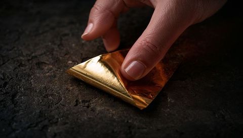 Closeup adult hand pressing gold foil on dark stone surface casting warm reflective sheen