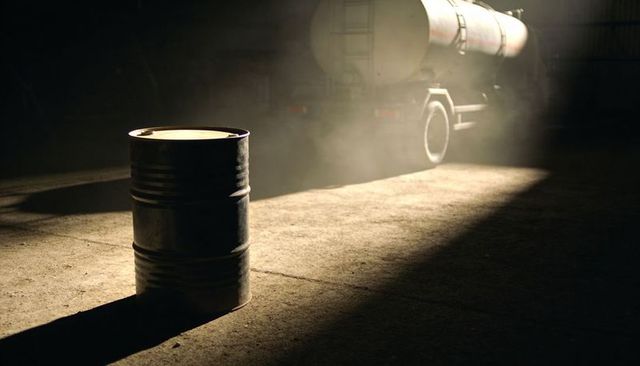 Industrial steel drum casting long shadow in spotlight with tanker truck silhouette moody