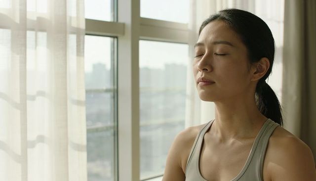 Asian woman practicing mindfulness and deep breathing by window with sheer curtains