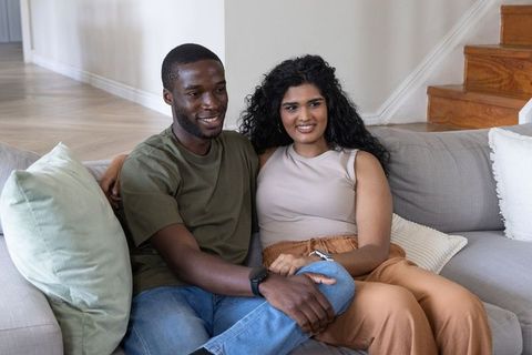 Diverse couple relaxing on modern sofa in cozy neutral living room, smiling and cuddling