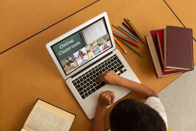 This image shows a child actively participating in an online class via a laptop at home. The screen displays a virtual classroom with multiple students and a teacher. Books and colored pencils indicate a study environment. Perfect for illustrating articles or blogs about e-learning, homeschooling, and the benefits of digital education, this image can also be used by educational technology companies and online learning platforms for promotional materials.