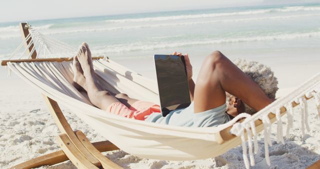 A woman is lying in a hammock on a sunny beach, leisurely using a tablet. Perfect for themes related to relaxation, technology in daily life, remote work scenarios, or digital lifestyles. Suitable for adverts or articles related to summer vacations, travel, and resort life.