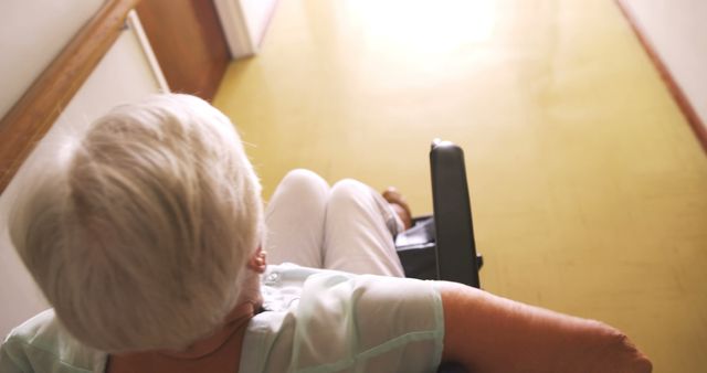 Elderly person with white hair navigating a wheelchair along a bright corridor with yellow flooring. The person's back is visible making their way towards a lighted area. Useful for topics related to elderly care, mobility aids, senior independence, nursing homes, and assisted living facilities.