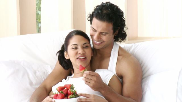 A loving couple enjoys eating strawberries while sitting comfortably in bed, radiating happiness and relaxation. Perfect for promoting romantic moments, intimacy, morning routines, and healthy living. Ideal for use in advertisements for fruits, mattress companies, or lifestyle blog articles focused on relationships and healthy snacks.