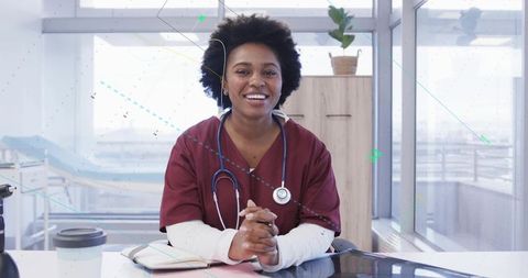 Smiling clinician in burgundy scrubs leaning at sunlit clinic desk with stethoscope and tablet
