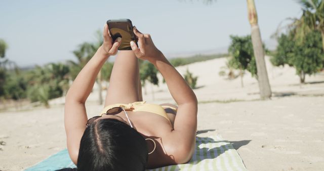 Woman lies on beach towel in bikini using smartphone under palm trees, ideal for summer vacation, relaxing lifestyle, or leisure activities themes.