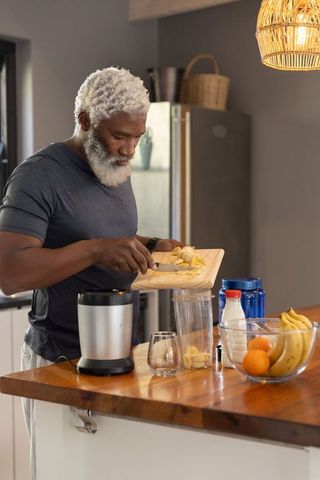 Senior african american man making banana smoothie at kitchen island, cozy healthy morning