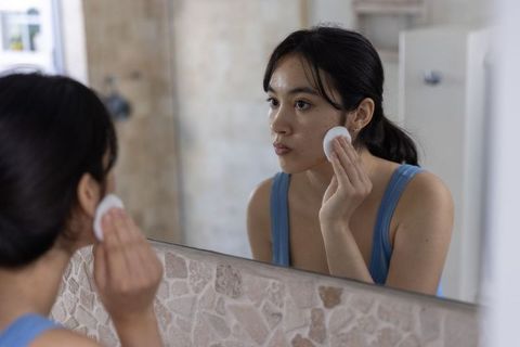Asian woman applying skincare with cotton pad at bathroom mirror morning routine