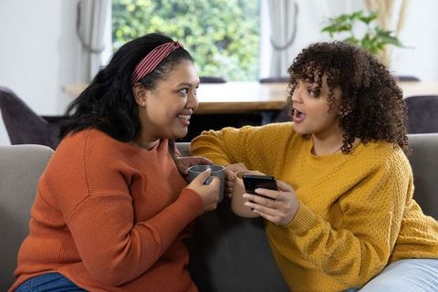 Two Friends Chatting on Sofa Holding Mugs and Smartphone Cozy Living Room Conversation