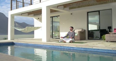Indian man meditating on yoga mat by pool at modern villa with hammock and mountain view