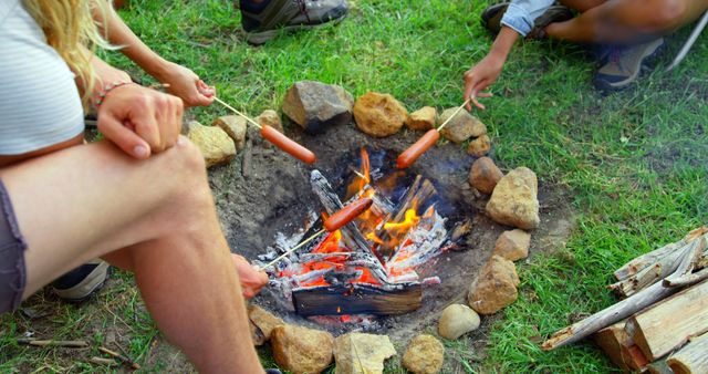 Group of friends enjoying outdoor adventure, roasting sausages over campfire in nature. Perfect for use in outdoor adventure advertisements, summer camp promotions, or content highlighting group fun activities.