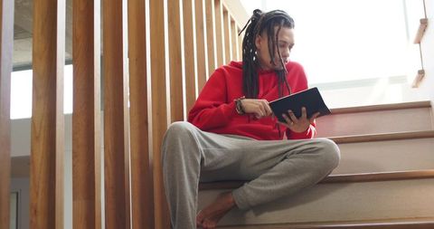 Man sitting on wooden staircase reading tablet wearing red hoodie and grey sweatpants