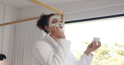 African american woman applying facial mask in white robe by sunny window for self care