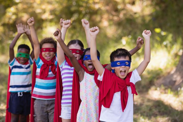 Group of diverse children wearing superhero costumes with red capes and masks, standing outdoors with arms raised in excitement. Ideal for themes of childhood, imagination, teamwork, and outdoor activities. Perfect for use in educational materials, advertisements for children's products, or community event promotions.