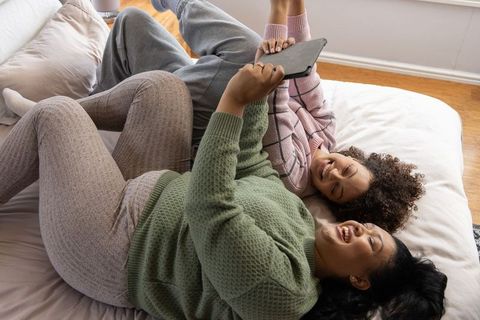 Diverse women laughing and scrolling tablet while lounging on bed in cozy sweater loungewear