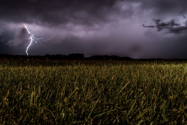 Dramatic scene capturing lightning bolt striking in distance across night sky with dark clouds gathering and green grass field in foreground. Useful for weather reports, blog articles on natural phenomena, environmental campaigns, or educational materials about thunderstorms.