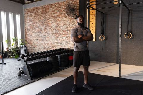 African american trainer standing with arms crossed in gym with dumbbells and rings