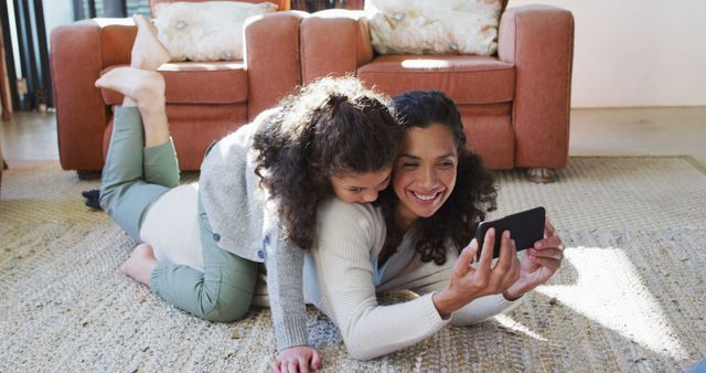 Happy biracial mother and daughter laying on the floor,having fun and taking selfie. domestic life and family leisure time concept.